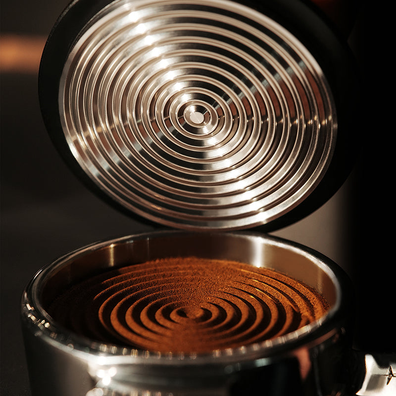 Close-up of the MHW-3BOMBER Knight Impact Tamper 58.35mm (Black) with its 4-spring internal design, displaying concentric patterns on finely ground coffee in a portafilter.