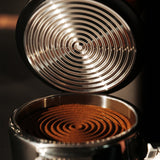 Close-up of the MHW-3BOMBER Knight Impact Tamper 58.35mm (Black) with its 4-spring internal design, displaying concentric patterns on finely ground coffee in a portafilter.