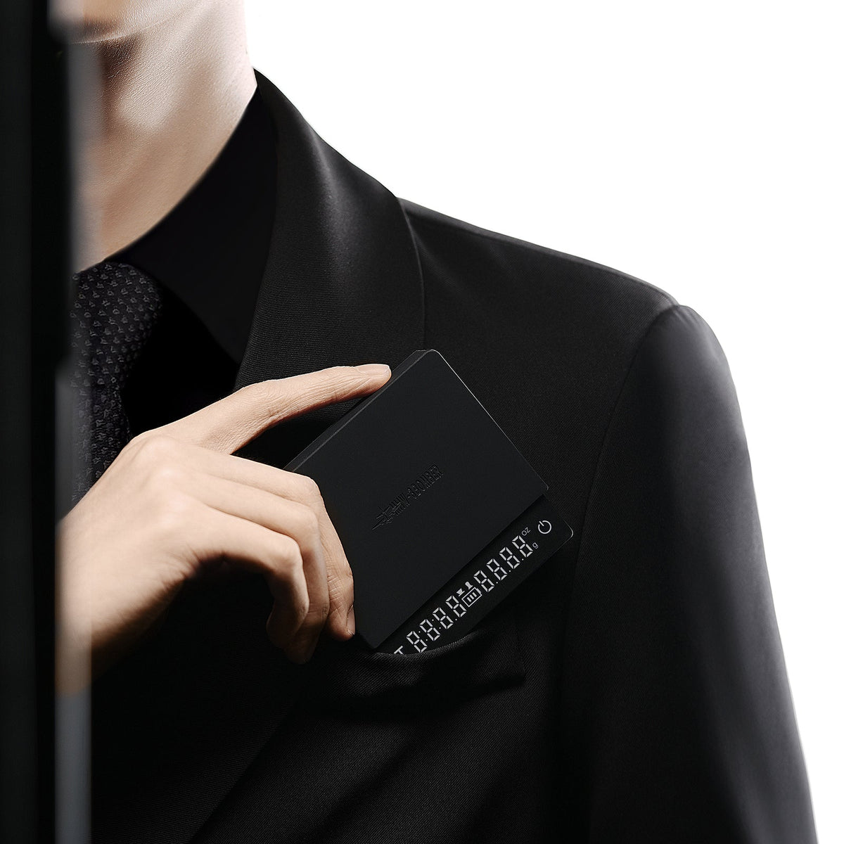 A person in a black suit and tie slips the MHW-3BOMBER Cube Coffee Scale 3.0 Micro—a sleek, black, rectangular device with numeric buttons—into their breast pocket against a white background.