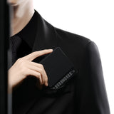 A person in a black suit and tie slips the MHW-3BOMBER Cube Coffee Scale 3.0 Micro—a sleek, black, rectangular device with numeric buttons—into their breast pocket against a white background.