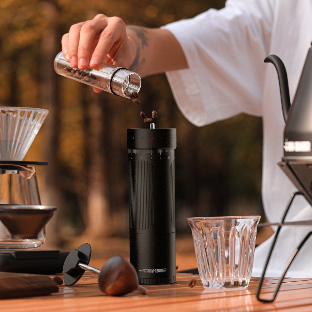 A person pours coffee beans into the MHW-3BOMBER Precision R3 Pro Manual Coffee Grinder (Black) on an outdoor table, next to a glass cup, pour-over dripper, and kettle with blurred trees in the background.