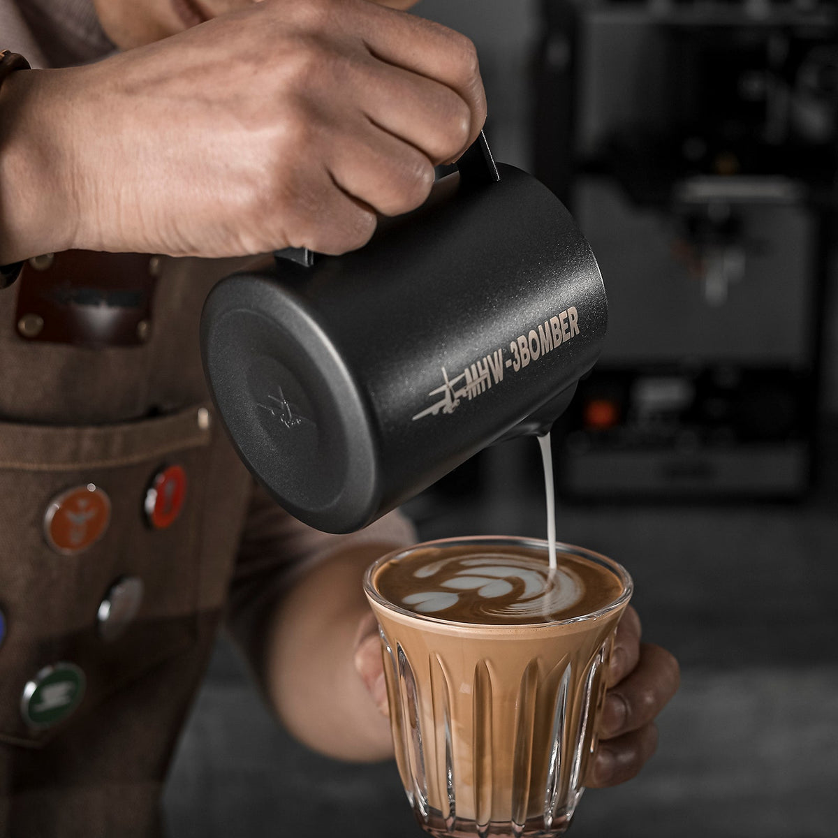 A barista pours steamed milk with the MHW-3BOMBER Latte Art Milk Pitcher 5.0 500ml (Matte Black), creating latte art, while colorful pins decorate their apron against a blurred background.