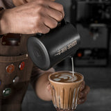 A barista pours steamed milk with the MHW-3BOMBER Latte Art Milk Pitcher 5.0 500ml (Matte Black), creating latte art, while colorful pins decorate their apron against a blurred background.