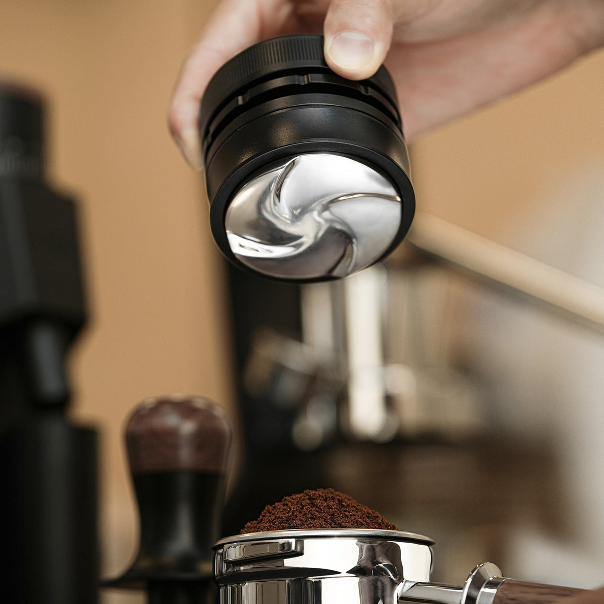 A hand holds the MHW-3BOMBER Coffee Gravity Distributor 58.35mm above a portafilter filled with ground coffee, ready for espresso preparation.