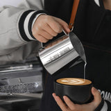 A person pours steamed milk from the MHW-3BOMBER Latte Art Milk Pitcher 5.0 500ml (Stainless Steel) into a black coffee cup, creating a latte art design on top.