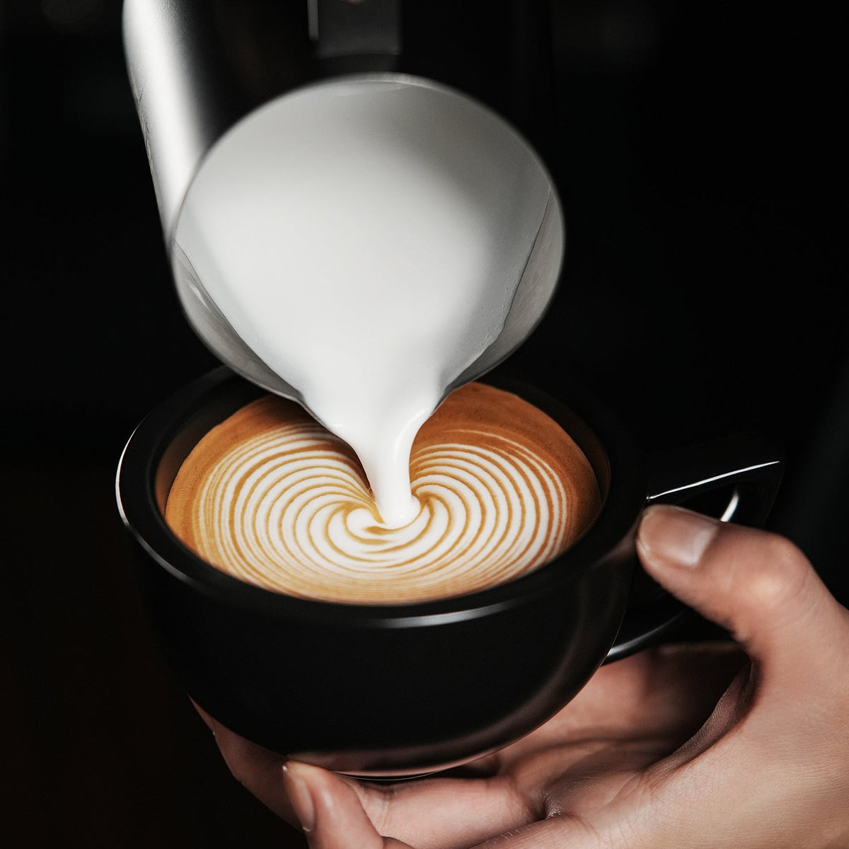 A hand holds a black cup of coffee as milk from the MHW-3BOMBER Latte Art Trio Bundle is poured in, creating intricate concentric circles on the surface.