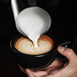A hand holds a black cup of coffee as milk from the MHW-3BOMBER Latte Art Trio Bundle is poured in, creating intricate concentric circles on the surface.