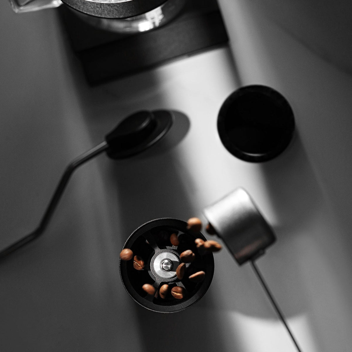 Overhead view of coffee beans falling into the MHW-3BOMBER Precision R3 Pro Manual Coffee Grinder (Black), with a scoop and kettle nearby on a white surface in dramatic lighting.