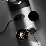 Overhead view of coffee beans falling into the MHW-3BOMBER Precision R3 Pro Manual Coffee Grinder (Black), with a scoop and kettle nearby on a white surface in dramatic lighting.
