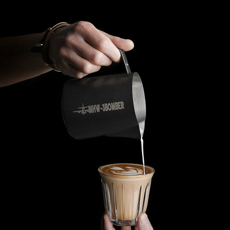 A hand pours milk from an MHW-3BOMBER Latte Art Milk Pitcher 5.0 500ml (Matte Black) into coffee, creating latte art with a crocodile-mouth spout against a black background.