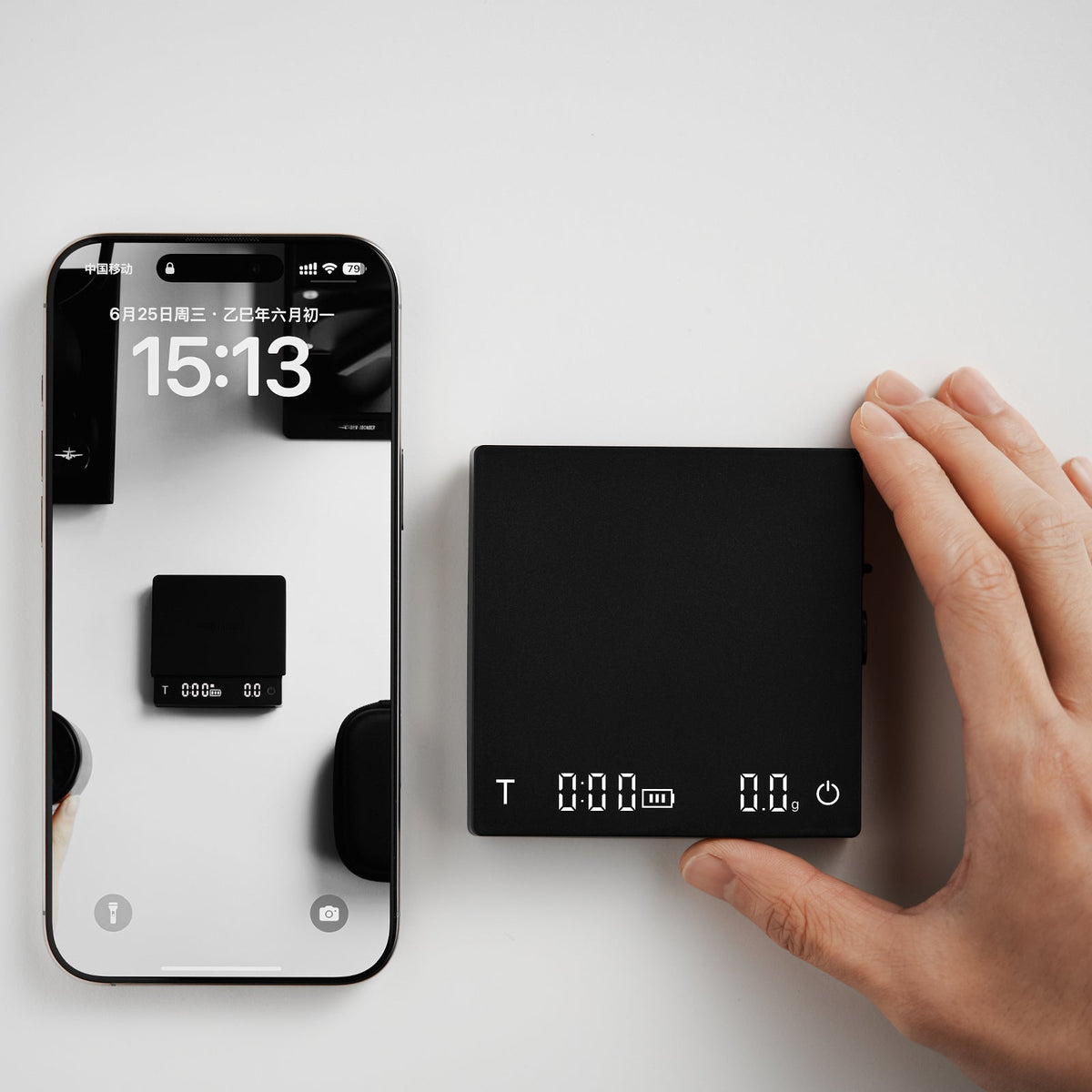 A hand presses the MHW-3BOMBER Cube Coffee Scale 3.0 Micro beside a smartphone displaying a clock and reflecting the device, both resting on a white surface.
