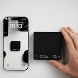 A hand presses the MHW-3BOMBER Cube Coffee Scale 3.0 Micro beside a smartphone displaying a clock and reflecting the device, both resting on a white surface.