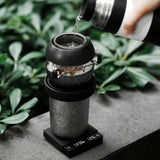 A hand pours hot water from a thermos into a coffee dripper set on a cup, which rests atop the MHW-3BOMBER Cube Coffee Scale 3.0 Micro, with green plants in the background.