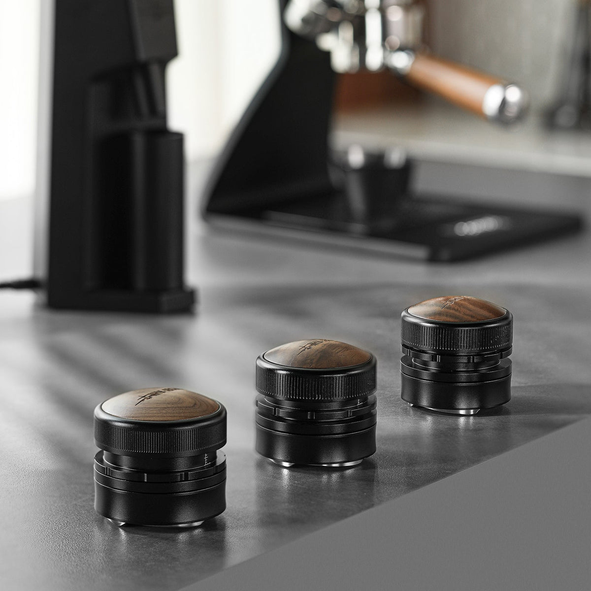 Three MHW-3BOMBER Coffee Gravity Distributor 58.35mm devices with wooden tops and cyclone bases are lined up on a gray counter, with a blurred espresso machine and portafilter in the background.