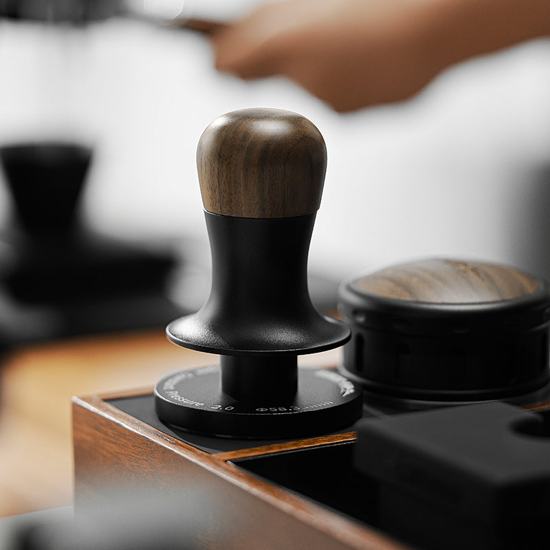 A close-up of the MHW-3BOMBER Flash Tamper 30lbs Constant Pressure Espresso Tamper 2.0 Sound Feedback with Ripple Base (Walnut) rests on a wooden tray, softly blurred alongside a hand and coffee tools.