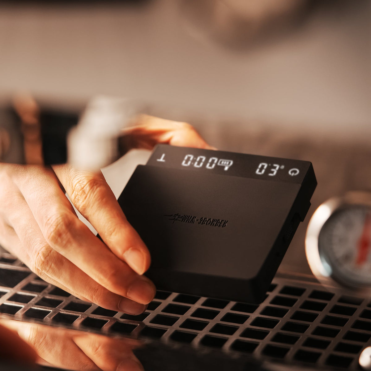 A hand holds the MHW-3BOMBER Cube Coffee Scale 3.0 Micro, its digital display showing numbers above a metal grate, while a blurred thermometer appears in the background.