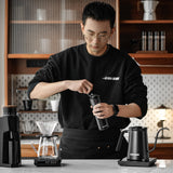 Someone grinds coffee beans in the kitchen with the MHW-3BOMBER Precision R3 Pro Manual Coffee Grinder (Black), alongside a pour-over setup and electric kettle on the counter, with shelves and kitchen items in the background.