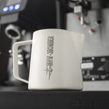 The MHW-3BOMBER Latte Art Milk Pitcher 5.0 500ml (White) sits on an espresso machine tray, featuring a plane graphic and “MHW-3BOMBER” branding, with blurred coffee equipment in the background.