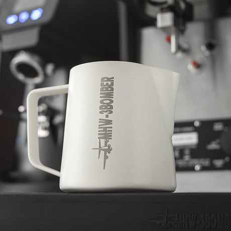 The MHW-3BOMBER Latte Art Milk Pitcher 5.0 500ml (White) sits on an espresso machine tray, featuring a plane graphic and “MHW-3BOMBER” branding, with blurred coffee equipment in the background.