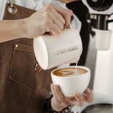 A person in a brown apron pours milk from the MHW-3BOMBER Latte Art Milk Pitcher 5.0 500ml (White) into a cup of coffee, creating beautiful latte art.