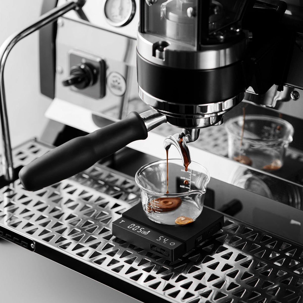 The MHW-3BOMBER Cube Coffee Scale 3.0 Micro precisely measures as an espresso machine brews coffee into a glass cup, with fresh espresso pouring from the portafilter.