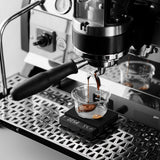 The MHW-3BOMBER Cube Coffee Scale 3.0 Micro precisely measures as an espresso machine brews coffee into a glass cup, with fresh espresso pouring from the portafilter.