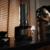 A sleek black coffee grinder sits by the MHW-3BOMBER X-press58 Portable Espresso Machine CM6954 (Black) and a neat stack of coffee pucks, while a hand holds a lid above them. Blinds and soft lighting are in the background.