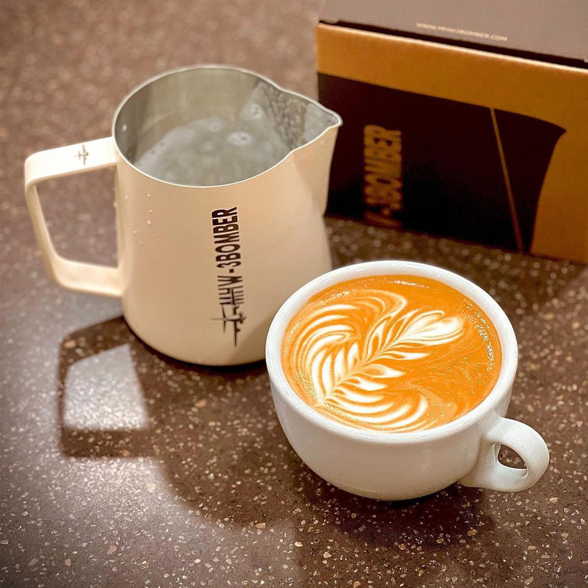 A latte with leaf art sits on a brown speckled surface next to the MHW-3BOMBER Latte Art Trio Bundle and a brown box.