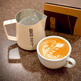 A latte with leaf art sits on a brown speckled surface next to the MHW-3BOMBER Latte Art Trio Bundle and a brown box.