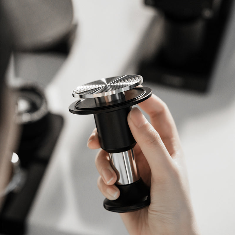 A hand holds the MHW-3BOMBER Knight Impact Tamper 58.35mm (Black) with its textured top and 4-spring design, set against a blurred backdrop of kitchen equipment.