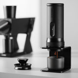 The MHW-3BOMBER X-press58 Portable Espresso Machine CM6954 (Black) dispenses coffee into a cup on its stand, with another coffee device and accessories blurred in a modern, minimalistic background.