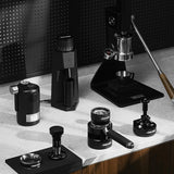 A countertop displays black coffee tools, including the MHW-3BOMBER YU Series Unibody Bottomless Portafilter 58mm, a manual espresso machine, grinder, tampers, and milk frother against a dark perforated backdrop.