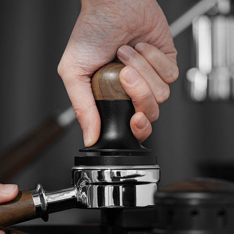 A close-up of a hand using the MHW-3BOMBER Flash Tamper 30lbs Constant Pressure Espresso Tamper 2.0 Sound Feedback with Ripple Base (Walnut) to press coffee grounds in a portafilter for espresso preparation.