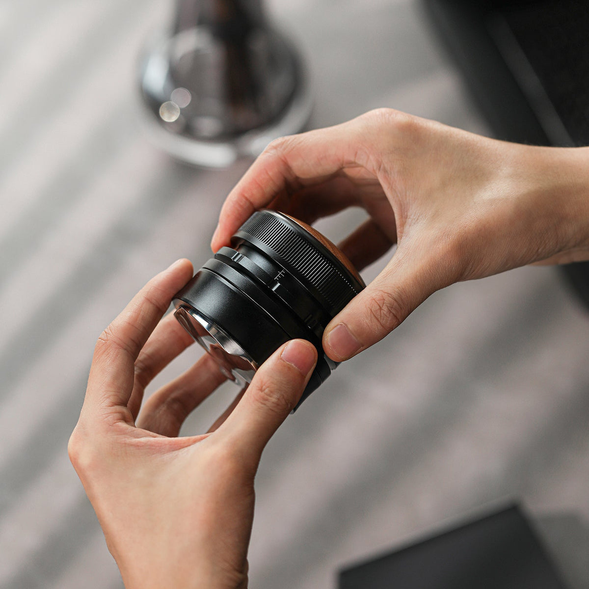 Two hands hold an MHW-3BOMBER Coffee Gravity Distributor 58.35mm above a blurred background, its small, black form featuring glass elements with cyclone-shaped reflections.