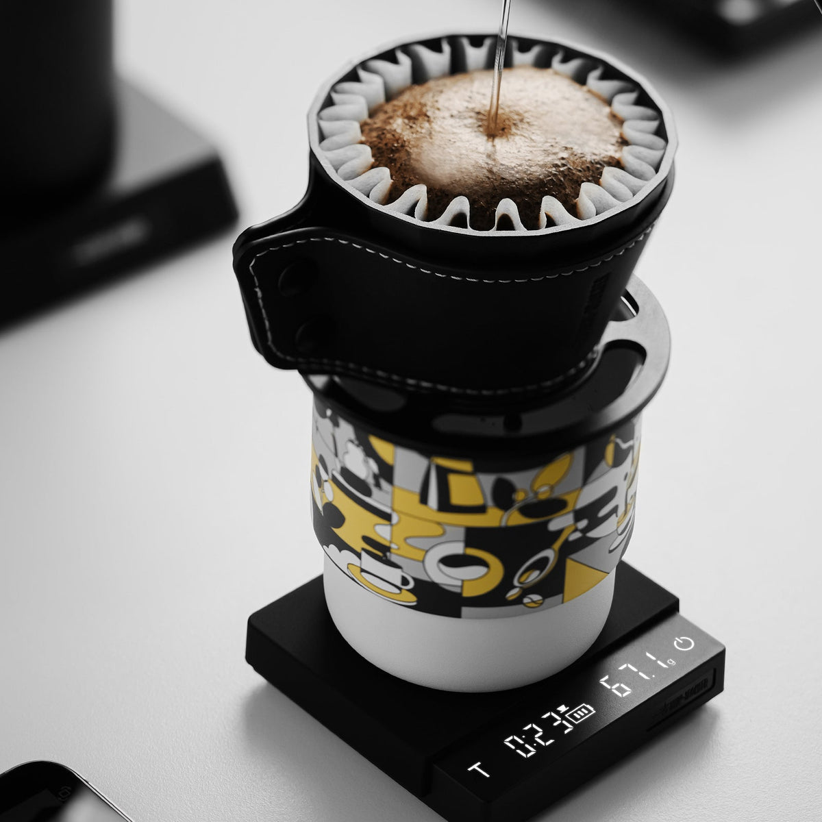 The MHW-3BOMBER Cube Coffee Scale 3.0 Micro from MHW-3BOMBER precisely tracks weight and time as coffee is brewed pour-over style into a patterned mug, with water poured over grounds in a filter atop the scale.