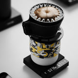 The MHW-3BOMBER Cube Coffee Scale 3.0 Micro from MHW-3BOMBER precisely tracks weight and time as coffee is brewed pour-over style into a patterned mug, with water poured over grounds in a filter atop the scale.