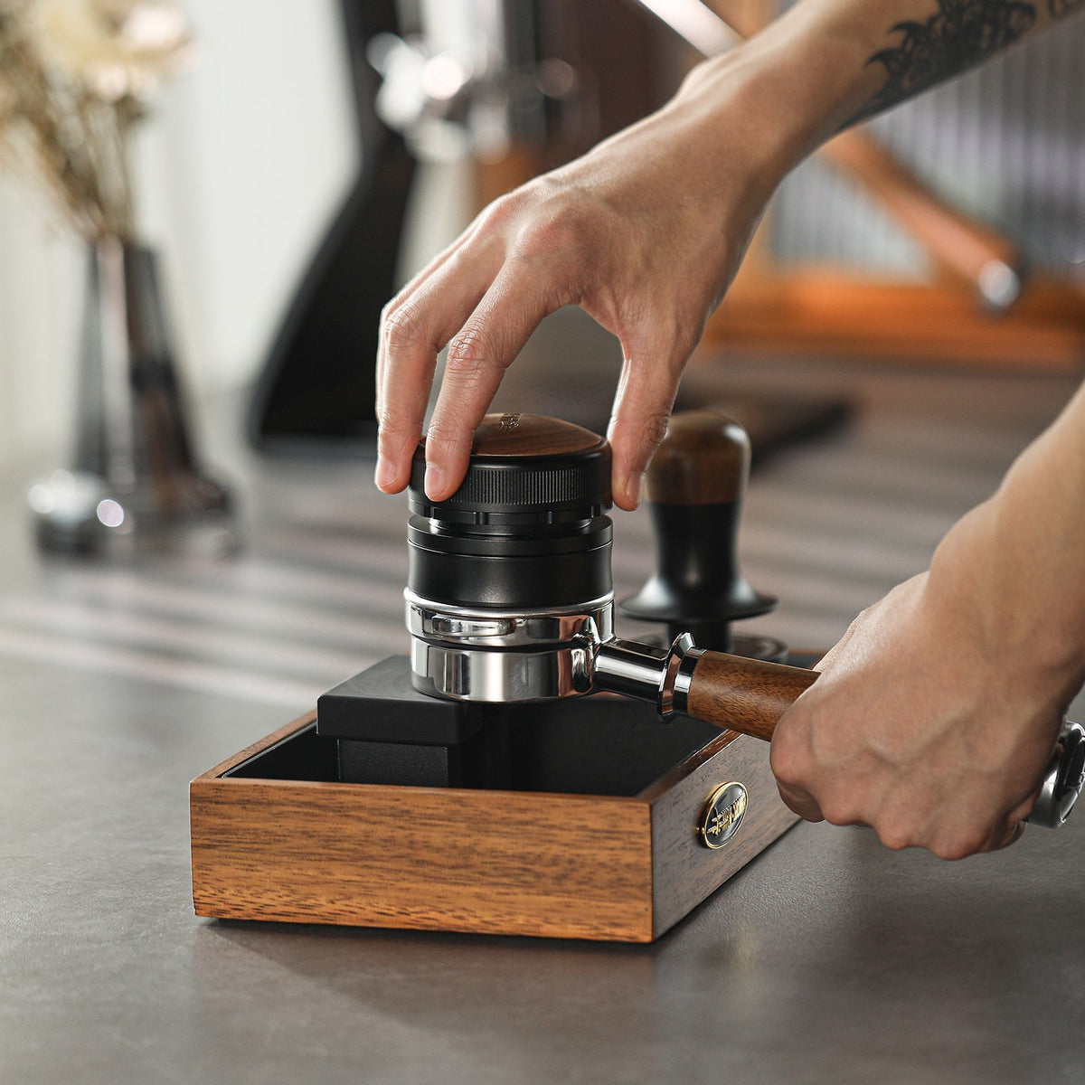 A person uses the MHW-3BOMBER Coffee Gravity Distributor 58.35mm on a portafilter in a wooden holder to prepare espresso on a kitchen counter.