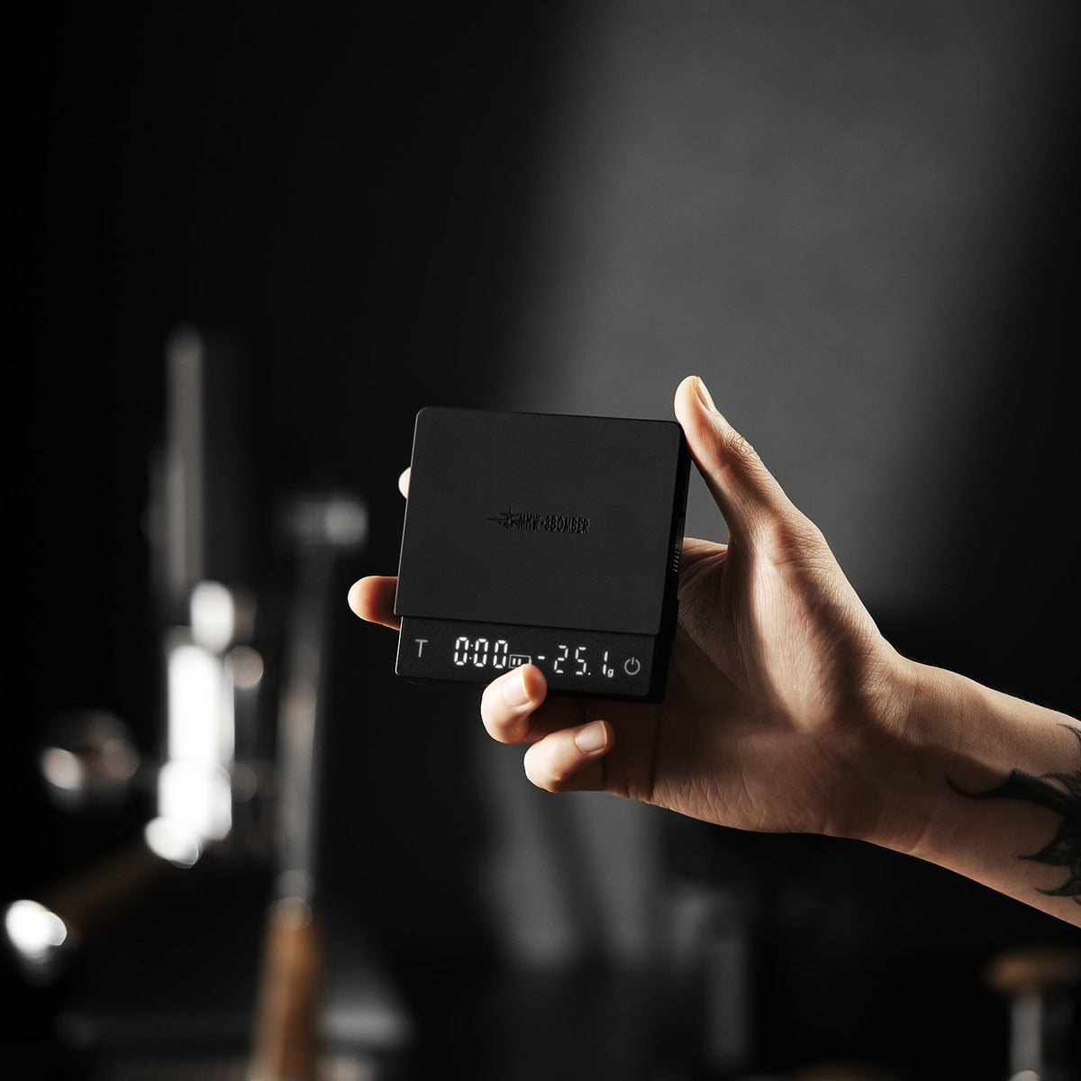 A hand holds the MHW-3BOMBER Cube Coffee Scale 3.0 Micro, its display showing “000 25.4”. The dark, blurry background draws attention to the precision of the scale.