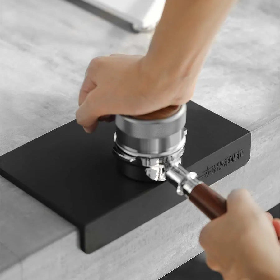 A person tamps coffee grounds into a portafilter on the MHW-3BOMBER Silicone Corner Tamping Mat SP5312B (Black) by MHW-3BOMBER on a gray countertop.