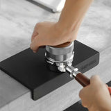 A person tamps coffee grounds into a portafilter on the MHW-3BOMBER Silicone Corner Tamping Mat SP5312B (Black) by MHW-3BOMBER on a gray countertop.
