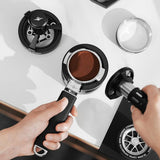 A person tamps ground coffee into an MHW-3BOMBER YU Series Unibody Bottomless Portafilter 58mm on a white countertop, with coffee tools and a distribution tool nearby.
