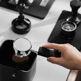 A hand knocks used coffee grounds from the MHW-3BOMBER YU Series Unibody Bottomless Portafilter 58mm into a black knock box, with coffee tools and a tamper on a white countertop.