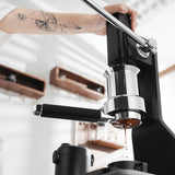 A hand uses a manual espresso machine with the MHW-3BOMBER YU Series Unibody Bottomless Portafilter 58mm, producing fresh espresso under bright light and shelves in the background.