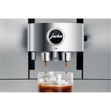 A Jura Z10 Aluminum White Super Automatic Coffee & Espresso Machine dispenses coffee over ice cubes into a glass, with droplets visibly falling from the Product Recognising Grinder’s spouts.