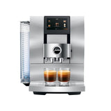 A Jura Z10 Aluminum White Super Automatic Hot Coffee & Espresso, Cold Brew, & Specialty Beverage Machine (model 15361 - Open Box) by Jura features a digital display and a sleek Swiss design. It is shown preparing two cups of espresso on its drip tray, with the water reservoir visible on the left side, highlighting its modern aesthetic and advanced features.