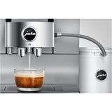 A Jura Z10 Aluminum White Super Automatic Hot Coffee & Espresso, Cold Brew, & Specialty Beverage Machine 15361 by Jura dispenses a creamy coffee into a glass cup, featuring its Product Recognising Grinder technology and a connected milk container beside it on a metallic countertop.