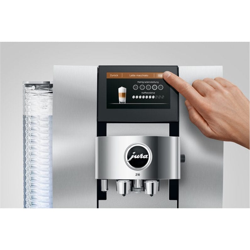 A hand selects "Latte macchiato" on the Jura Z10 Aluminum White Super Automatic Hot Coffee & Espresso, Cold Brew, & Specialty Beverage Machine 15361's digital touchscreen, with clear cups stacked on the left and its Product Recognising Grinder ensuring a perfect brew.