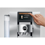 A hand selects "Latte macchiato" on the Jura Z10 Aluminum White Super Automatic Hot Coffee & Espresso, Cold Brew, & Specialty Beverage Machine 15361's digital touchscreen, with clear cups stacked on the left and its Product Recognising Grinder ensuring a perfect brew.