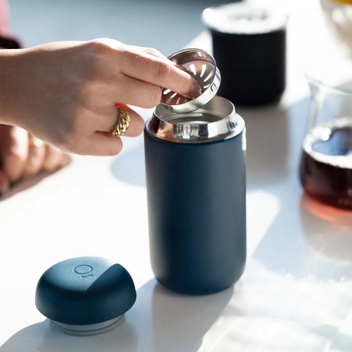 A hand places a metal infuser into a Fellow Carter Move Mug 16oz (Stone Blue) with a ceramic interior on a white surface. The mug's cap is nearby, and a glass carafe with dark liquid is in the background.