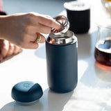 A hand places a metal infuser into a Fellow Carter Move Mug 16oz (Stone Blue) with a ceramic interior on a white surface. The mug's cap is nearby, and a glass carafe with dark liquid is in the background.